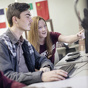 two students working at a computer together with flags from around the world visible
                     on the wall behind them