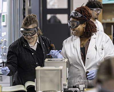 Two students wearing safety goggles and gloves working in a science lab.
