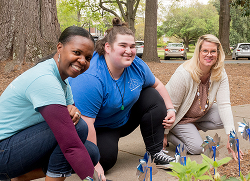 Two social work professors and a student smile while setting up pinwheels in the garden
                        outside Bancroft Hall
