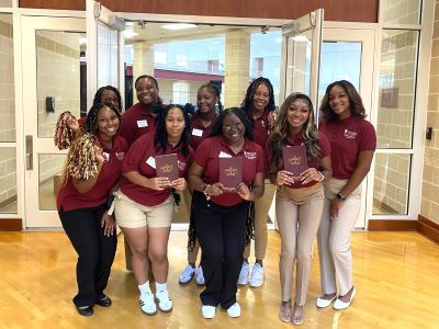 Members of the Student Alumni Council in matching garnet shirts holding up the convocation
                        program and pompoms. They are standing inside the West Center near the gymnasium