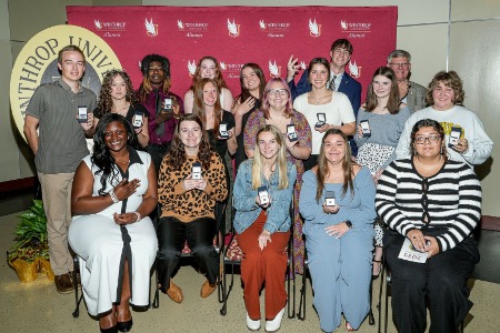 A group of students holding up their hands to show their Winthrop class rings in front
                        of a logo backdrop