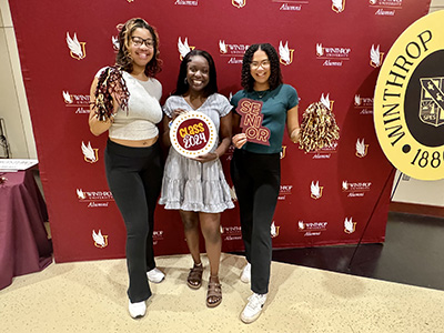 three students standing in front of the Winthrop logo holding pompoms and a Class
                        of 2024 sign