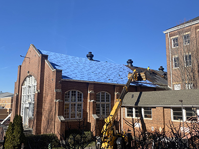 Blue sky with brick building in forefront. Yellow construction equipment pulling tile
                        from roof