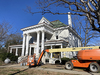 Large white house with porch columns in front of a blue sky. Orange construction truck
                        in front of it