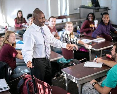 a professor walking between desks while talking to smiling students in class