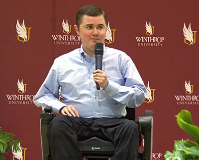 a student in professional attire holding a microphone in front of a wall with the
                        winthrop logo