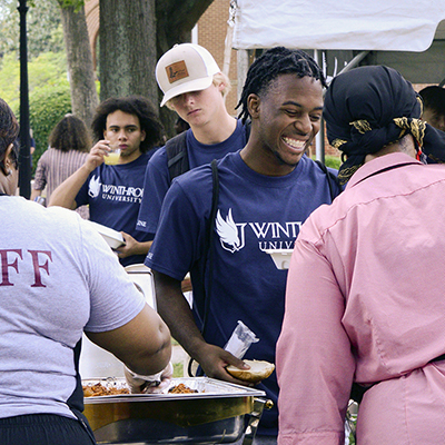 A student in a blue Winthrop University tshirt smiles while getting food at a convocation
                           community picnic