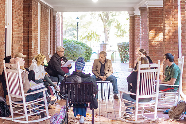 a professor and students holding class outside on a pretty day. They have set up chairs
                  in a circle on a historic brick porch