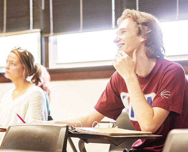 two students sitting at desks in the classroom and smiling at something the professor
                        just said