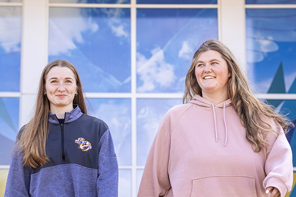 two undergraduate students walking outside with one of the student center window murals
                  behind them