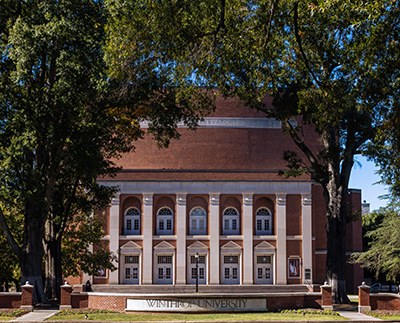 a photograph of the Winthrop University sign with Byrnes Auditorium in the background.
                        It is a sunny spring day and there are flowers blooming beside the sign