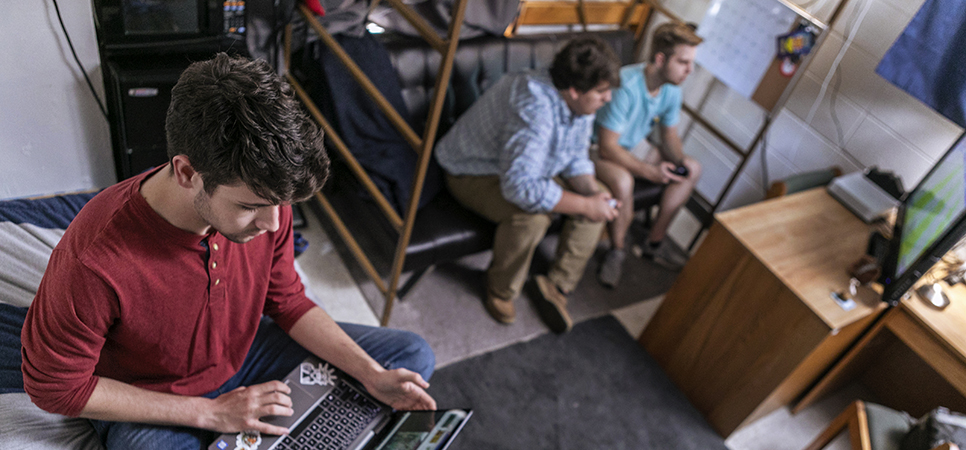 a student working on his laptop in his dorm room with a mini fridge and microwave
                                    visible behind him