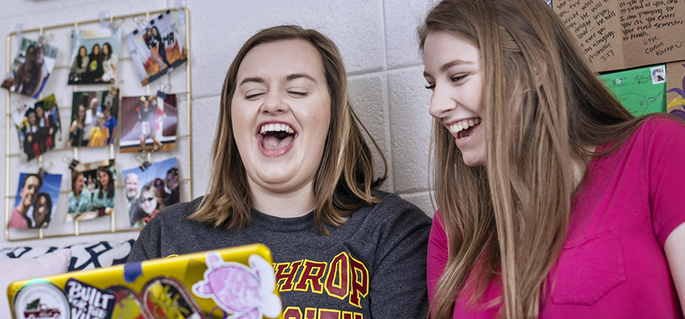 two roommates sitting together in their room and laughing at something they're watching
                                    on a laptop