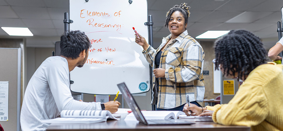 a tutor explaining a concept on a whiteboard to two students in the academic success
                                    center