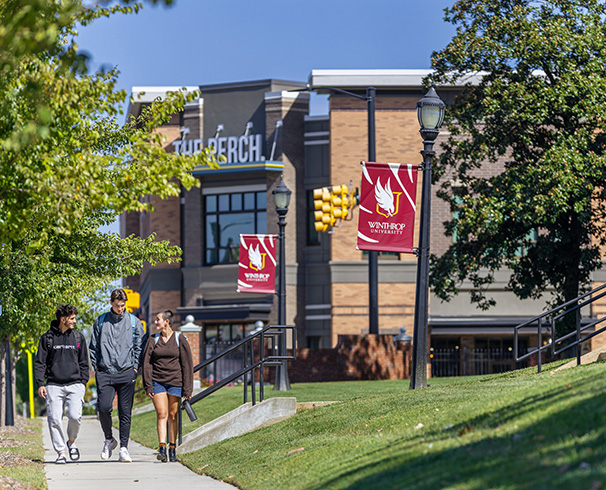 three students walking down the sidewalk near campus with Winthrop banners on the
                        lampposts and local restaurants visible in the background