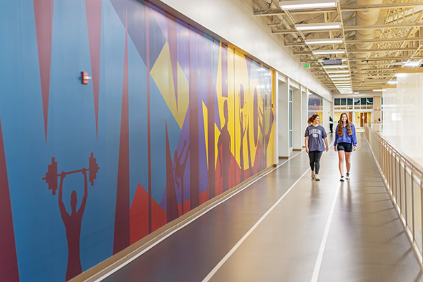 two students walking around the indoor track in the west center beside a colorful
                  Winthrop mural