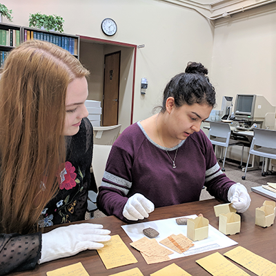 Two women wearing gloves and inspecting pages labels and clay pieces