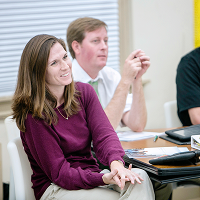 Woman in a purple sweater sitting at a classroom table and smiling at someone off-screen