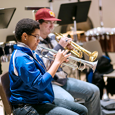 Two people playing trumpets with drums and music stands in the background